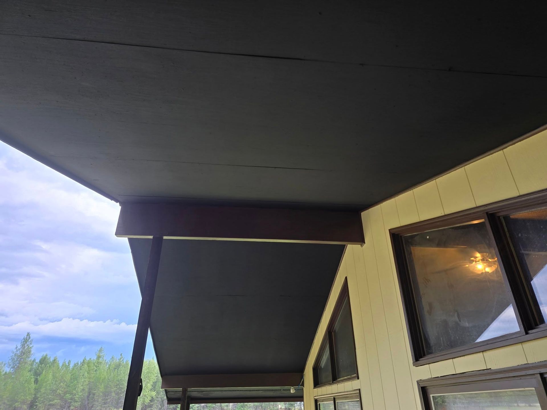Black painted ceiling of a porch. Brown wooden beam, beige exterior wall with windows. Trees in background.