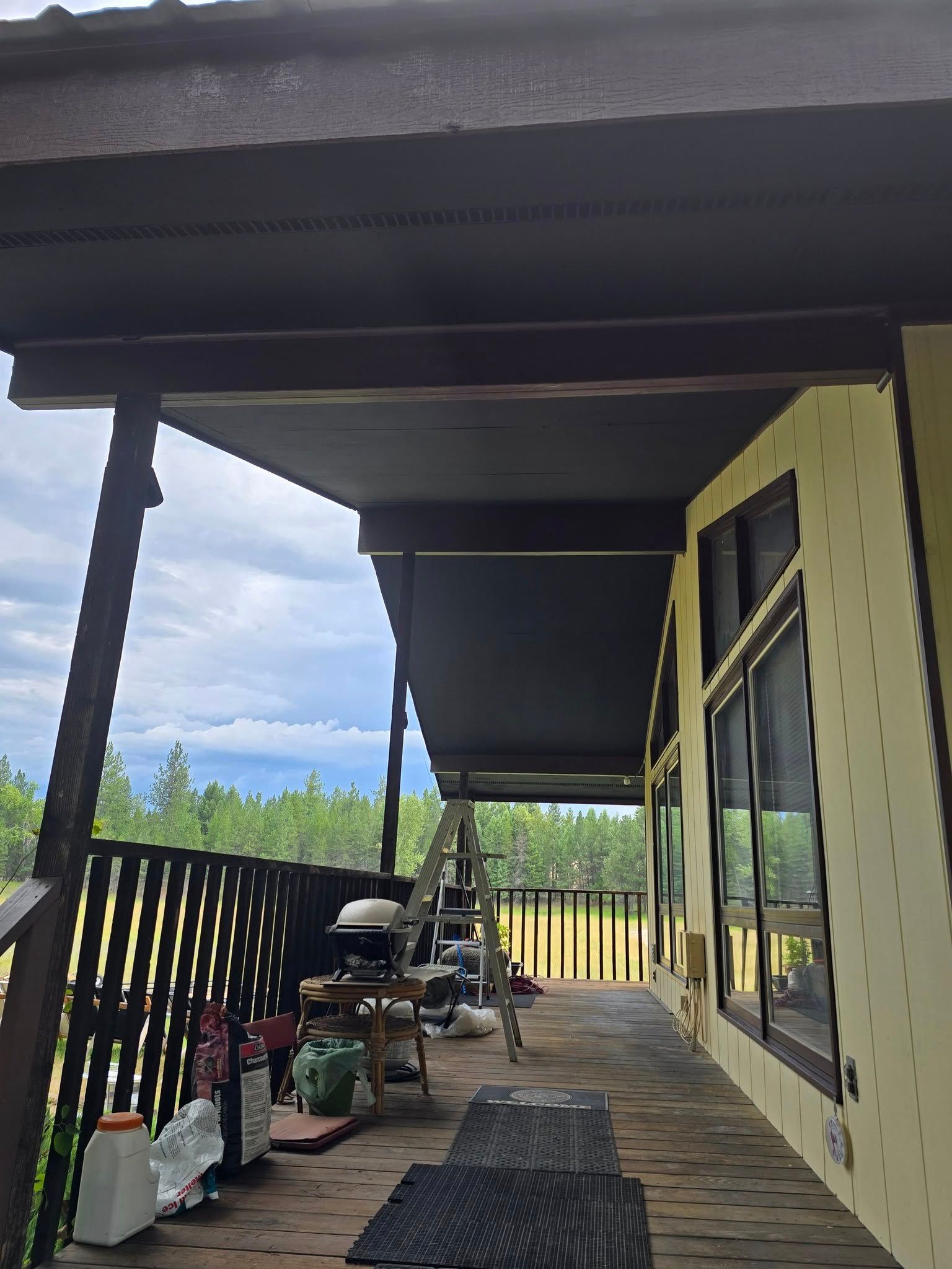 A covered porch with dark ceiling. Brown beams and railings with a view of a wooded area and cloudy sky.