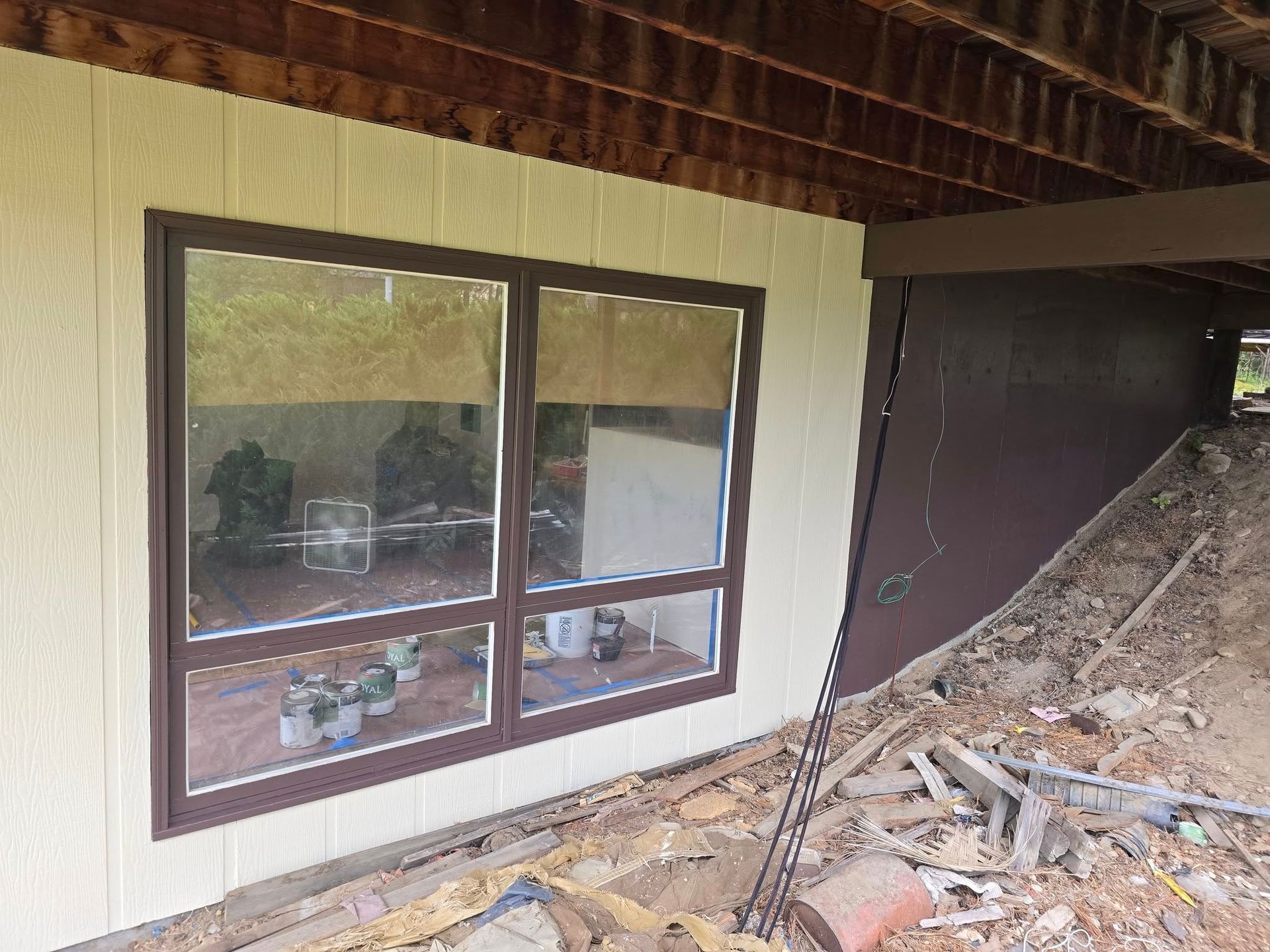 Brown-framed window on a building with beige siding, looking into a room with potted plants.