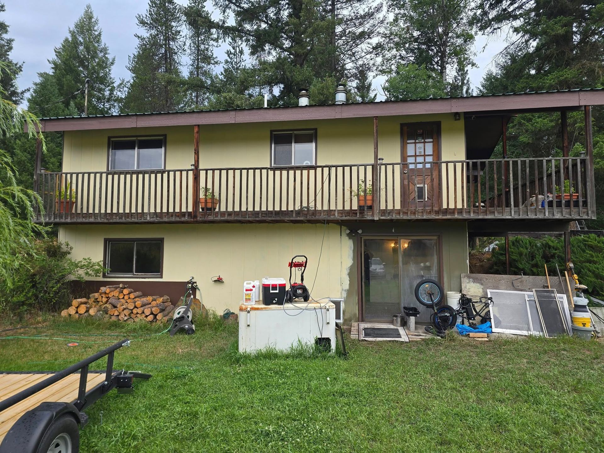 Two-story yellow house with balcony and wooden railing. Trailer and various items sit on the lawn.