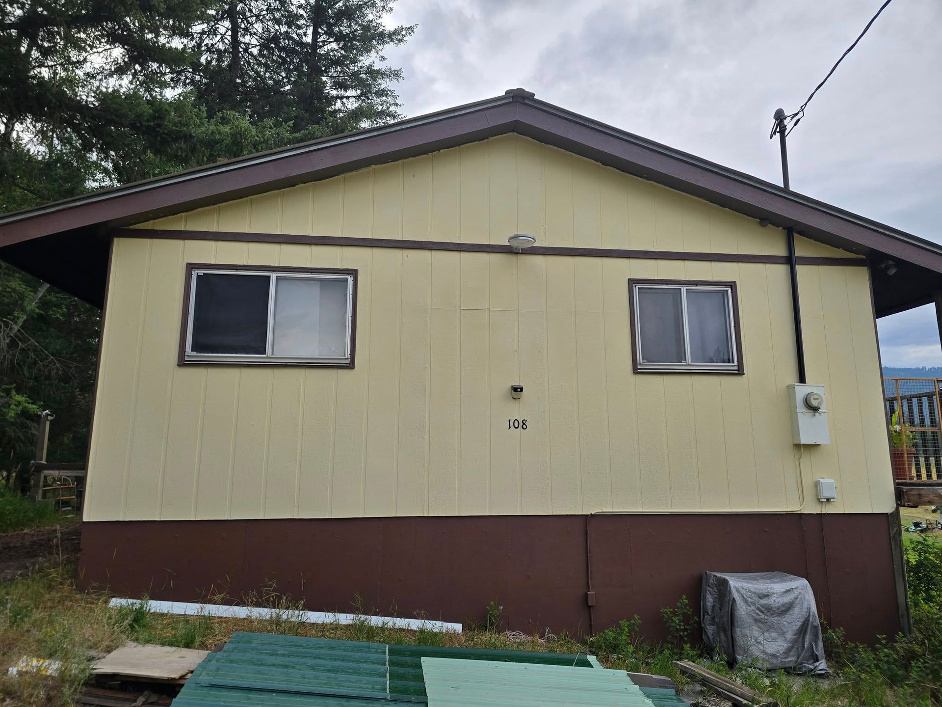Yellow and brown one-story building with two windows. Dark roof and trim. Exterior electrical box visible.