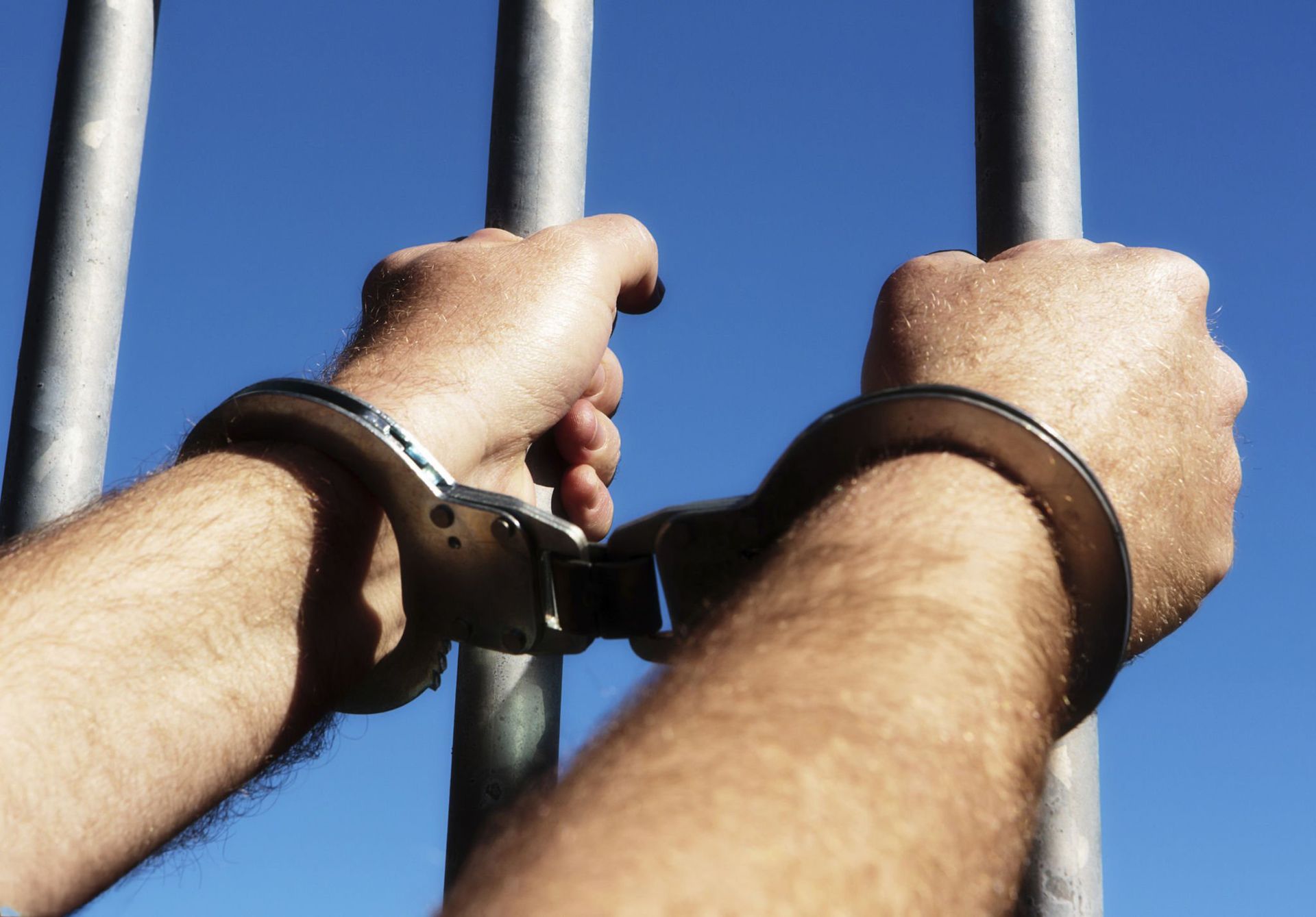 Hands in handcuffs gripping jail bars against bright blue sky. Hands in handcuffs gripping jail bars against bright blue sky.