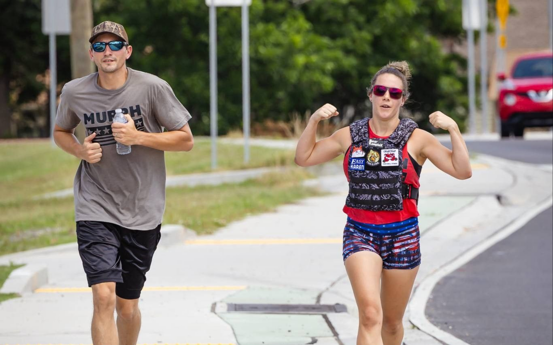 Two people run on a sidewalk; one wears a weight vest. Bright, outdoor setting.