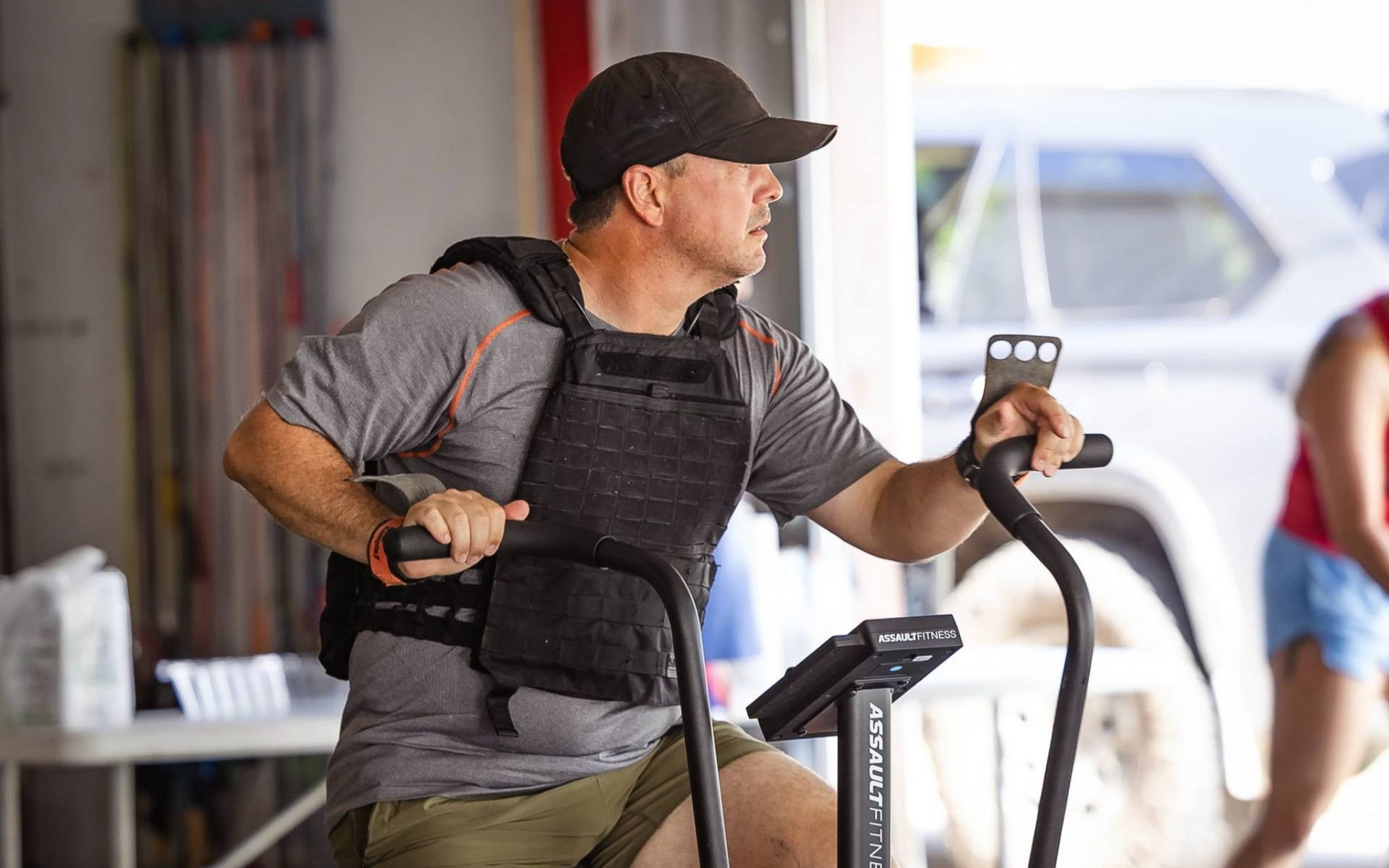 Man in gray shirt and baseball cap, wearing a weighted vest, exercising on an air bike indoors.