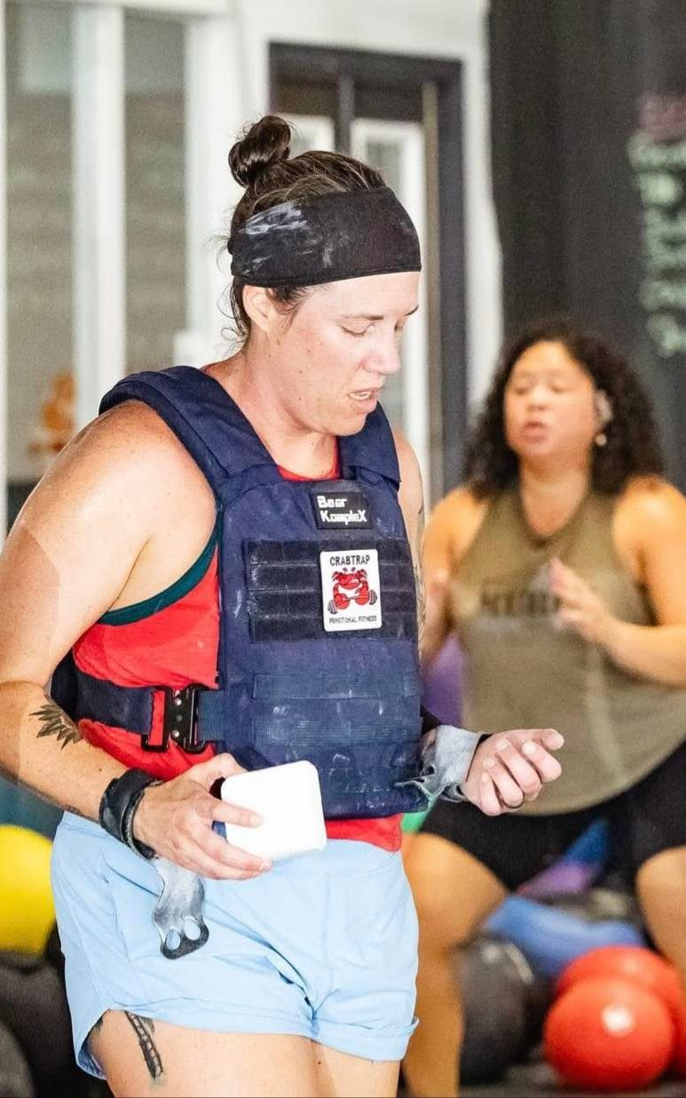 Woman in weight vest at gym, holding chalk, while another woman squats in background.