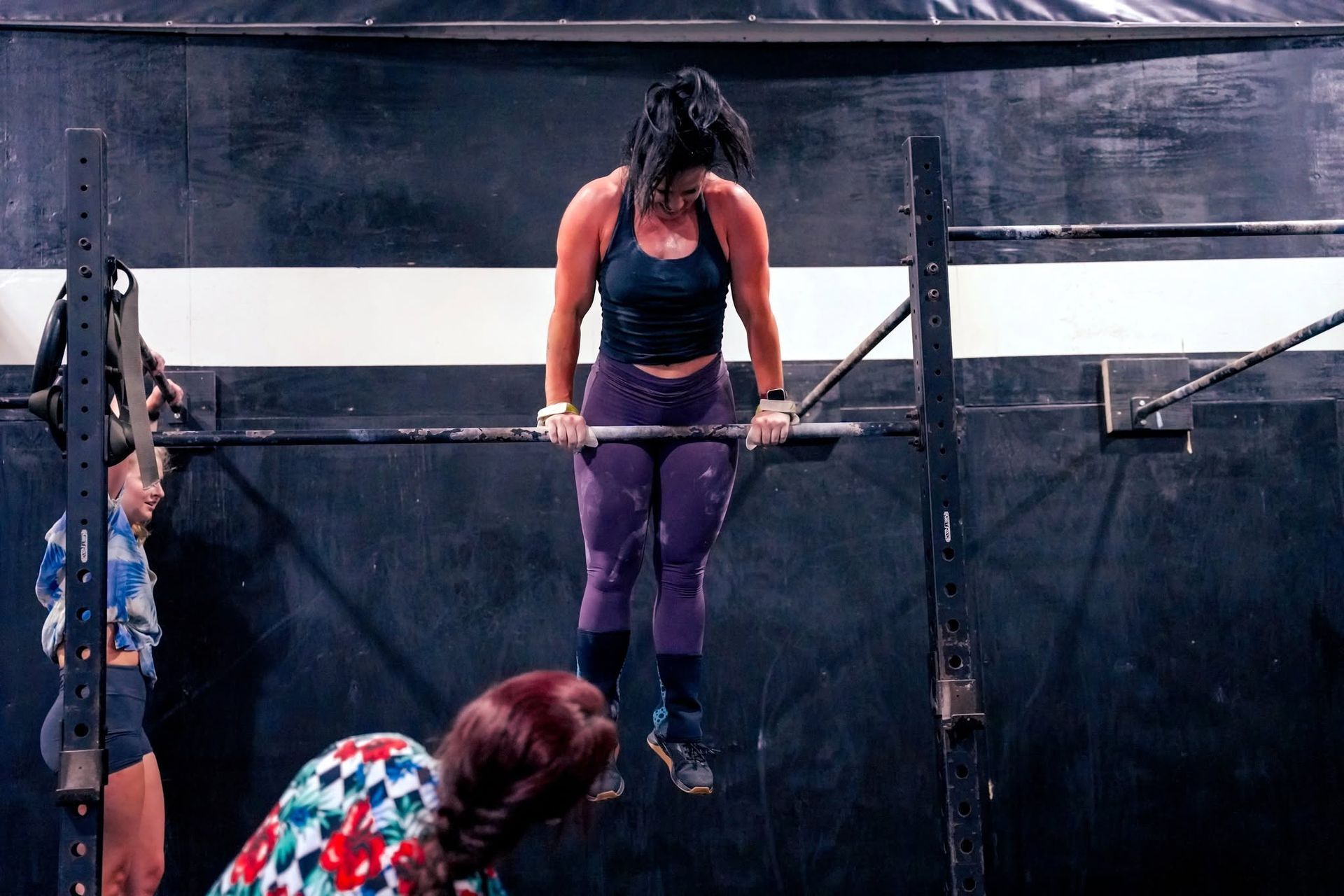 Woman hanging from a horizontal bar in an outdoor obstacle course, wearing purple leggings and a black tank top.