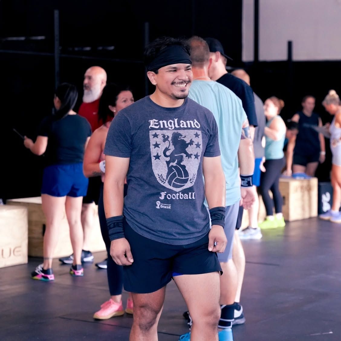 Smiling athlete in a gray T-shirt and black shorts stands in a gym with other people in the background