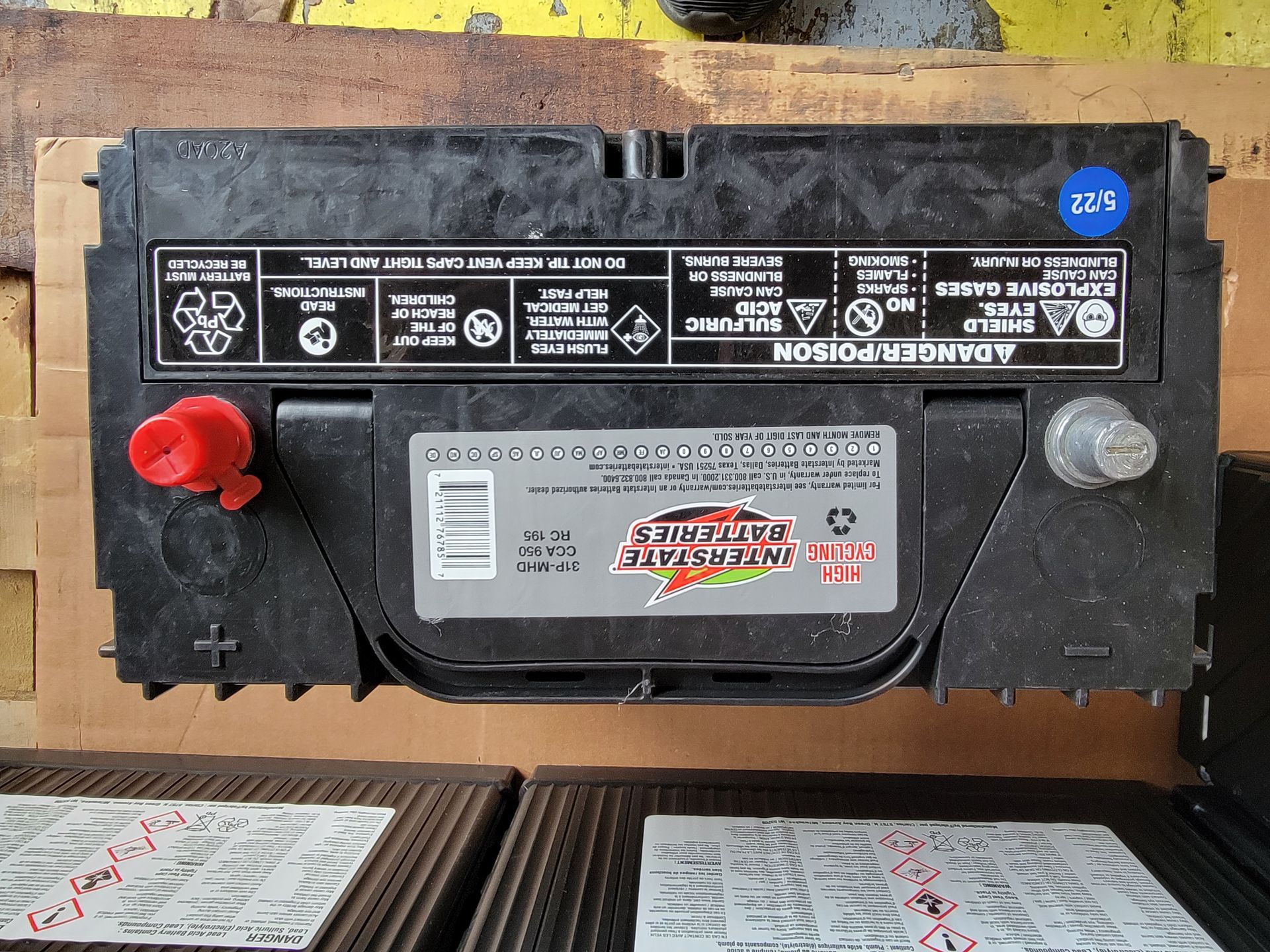 Black car battery with red and silver terminals, warning labels.