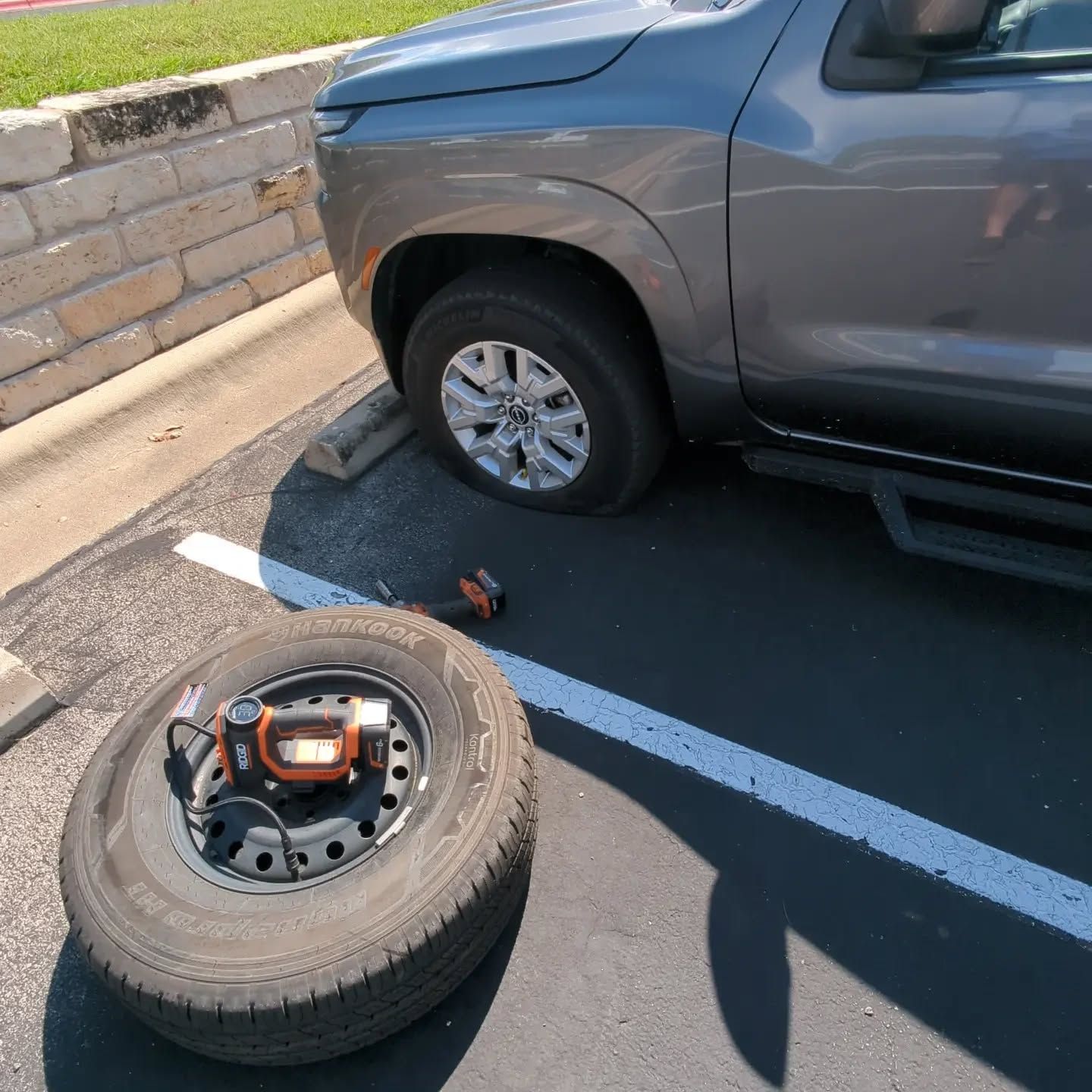 Flat tire on gray truck; spare tire on the ground with a tire inflator in a parking lot.
