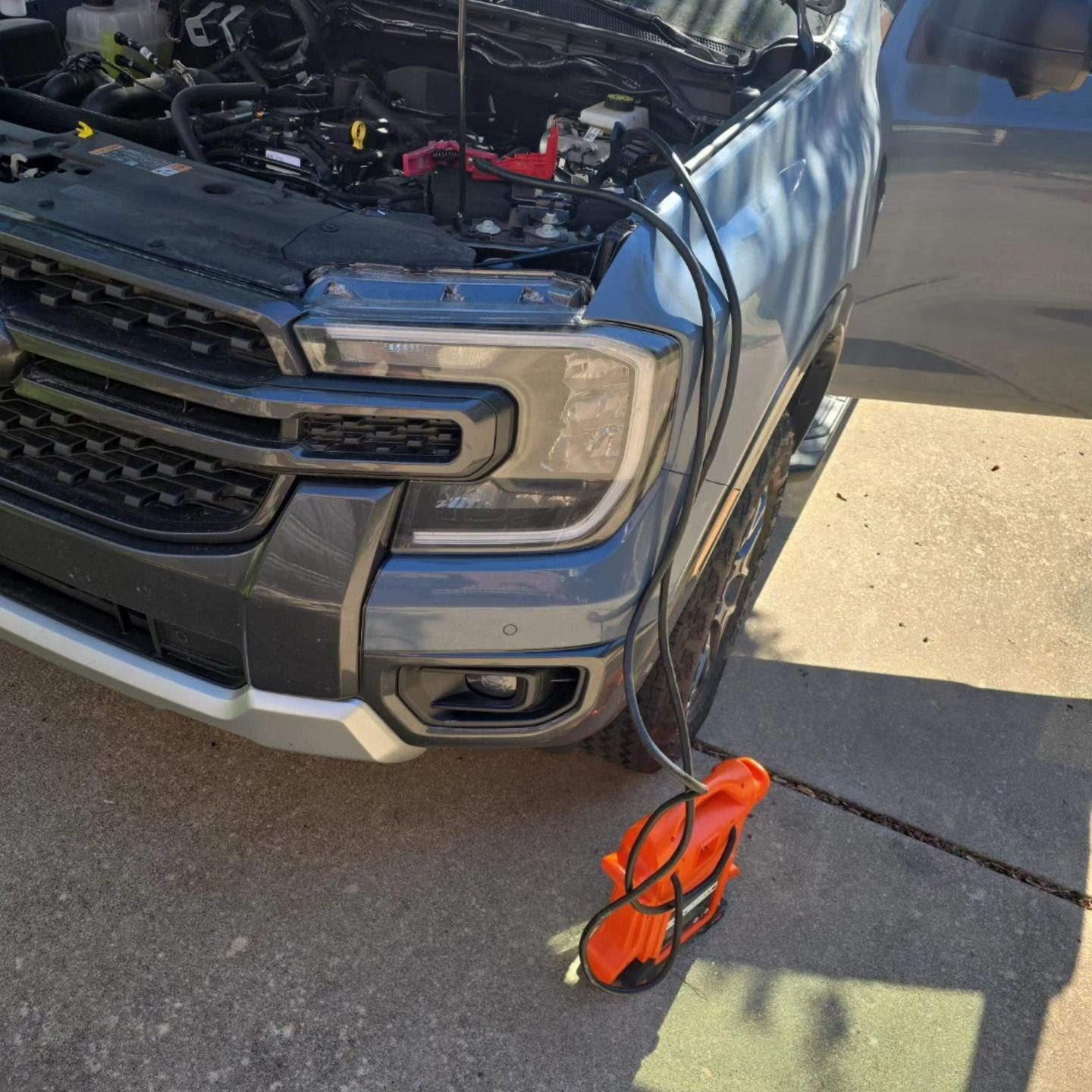 A gray car with its hood open, a black and orange blower nearby, outside on a driveway.