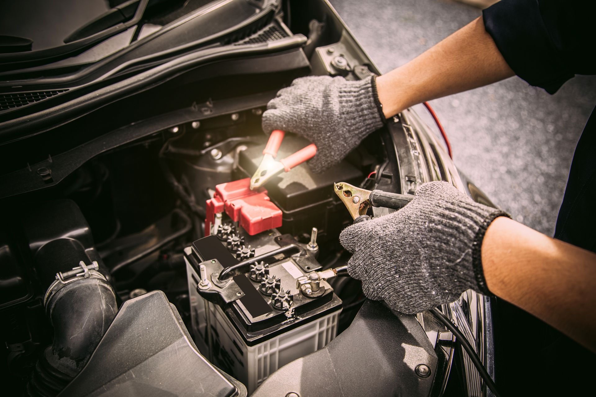 Hands in gloves connecting jumper cables to a car battery in engine bay.