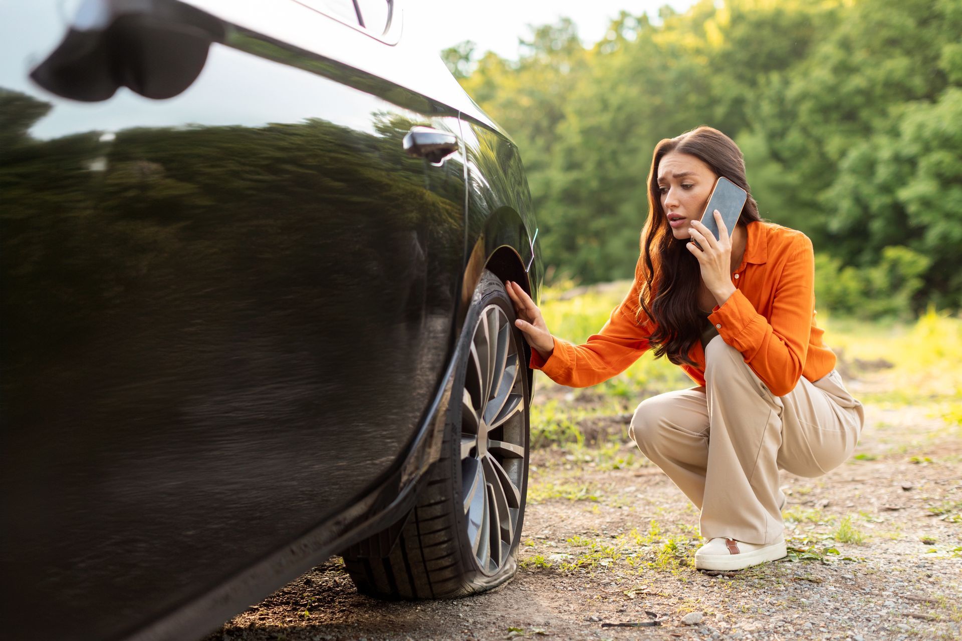 Woman with flat tire, crouching near black car, on phone, with concerned expression.