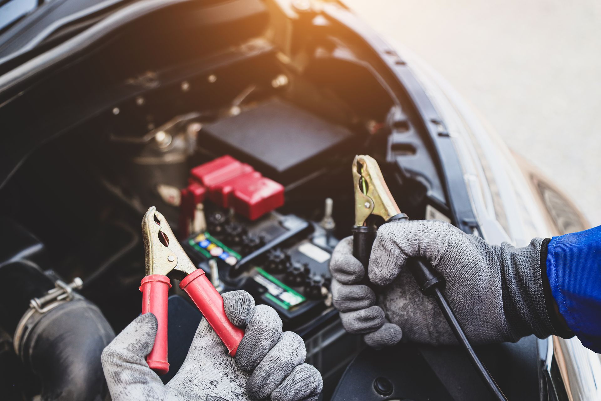 Hands in gloves connecting jumper cables to a car battery in an engine bay.