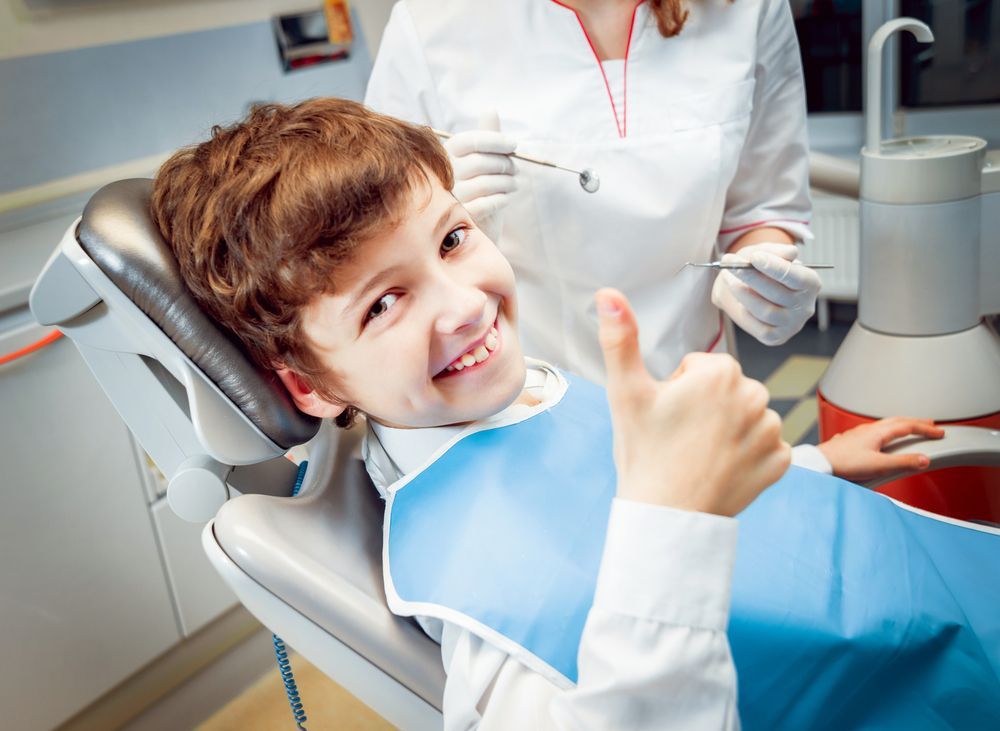 Boy in dentist chair gives thumbs-up, smiling. Dentist in white coat holds tools. Bright dental office.