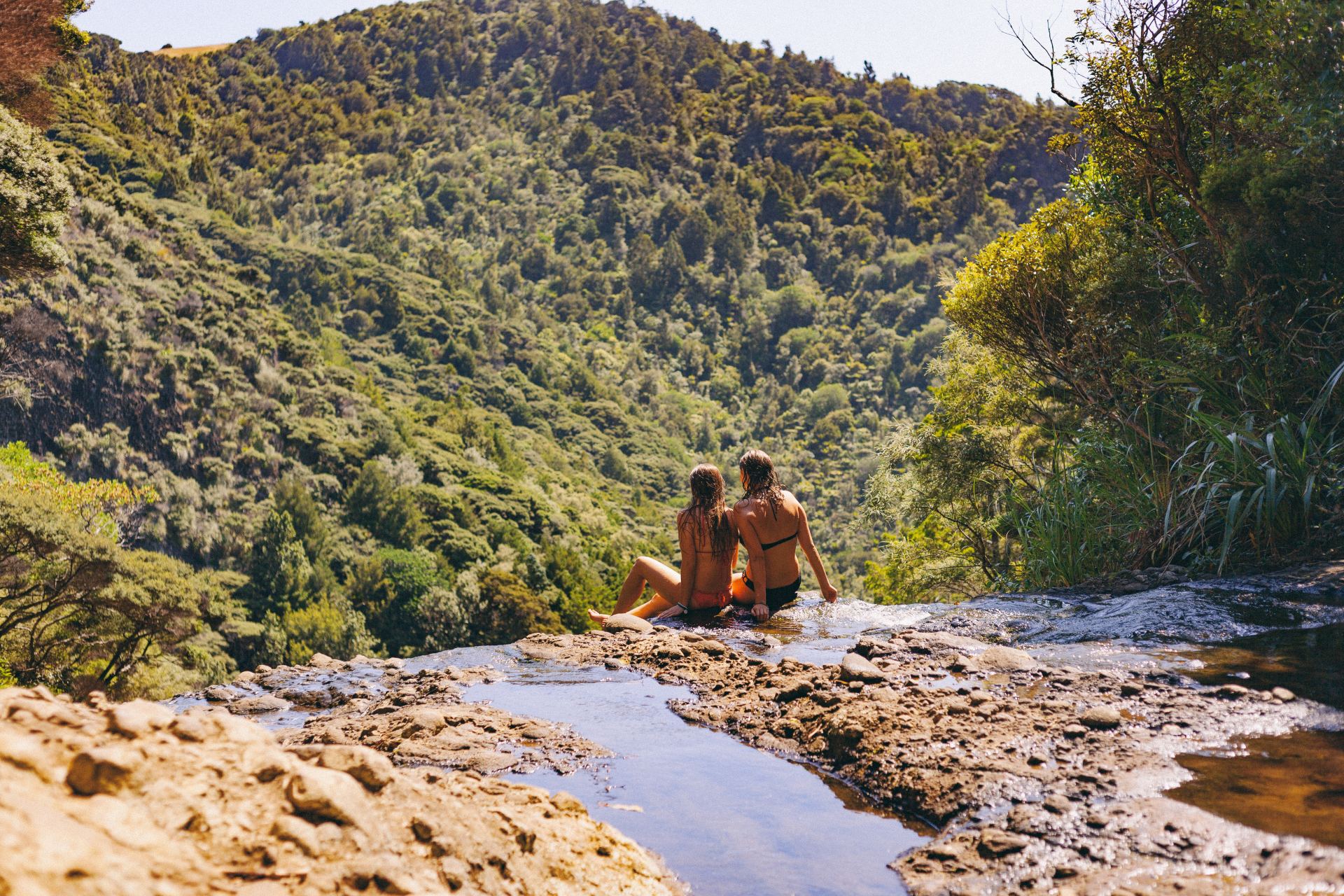 Imagem de duas  mulheres de biquíni sentadas nas pedras observando a cachoeira em Piha, Nova Zelândia