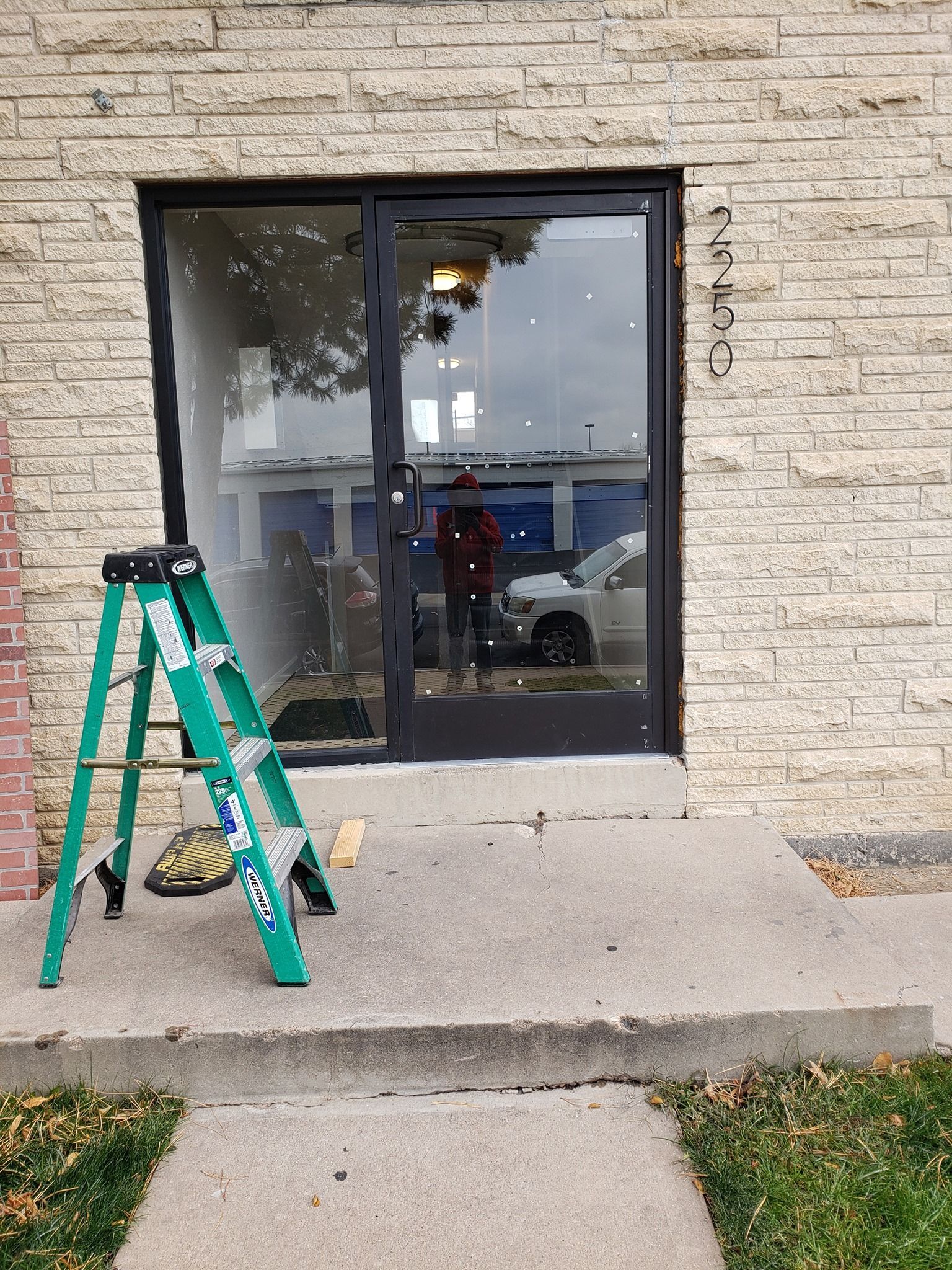Green ladder at a building entrance with a glass door, concrete steps, and a person reflected in the glass.