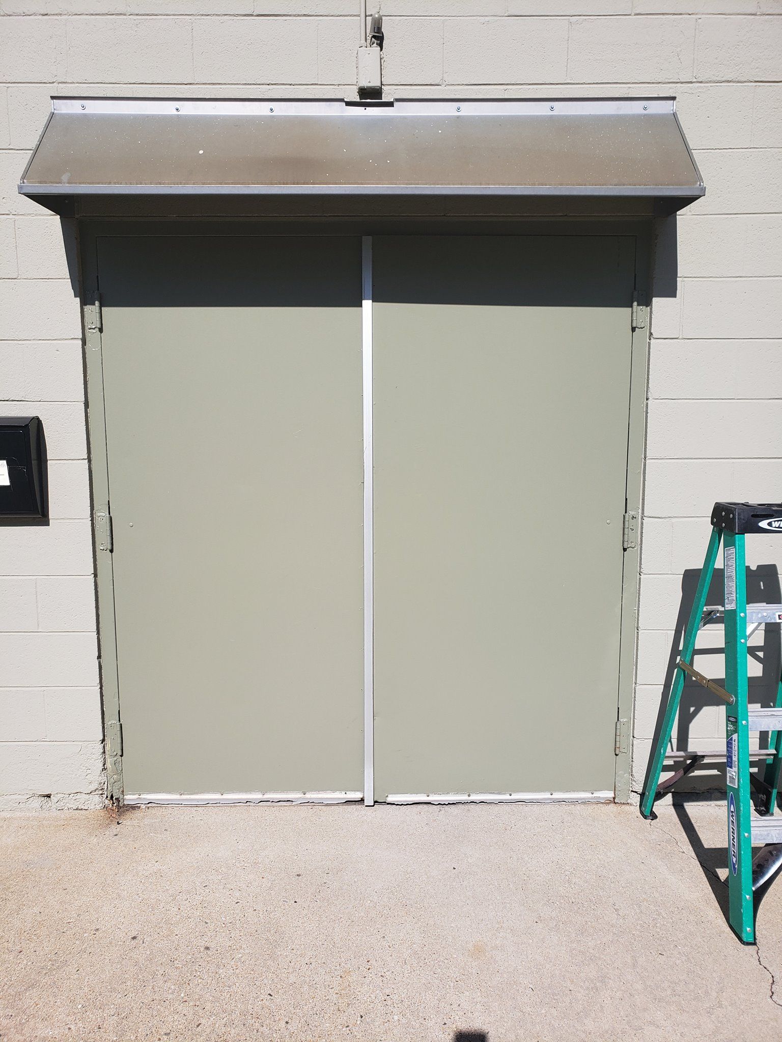 Green double doors with a silver frame under a small awning, with a green ladder on the right.