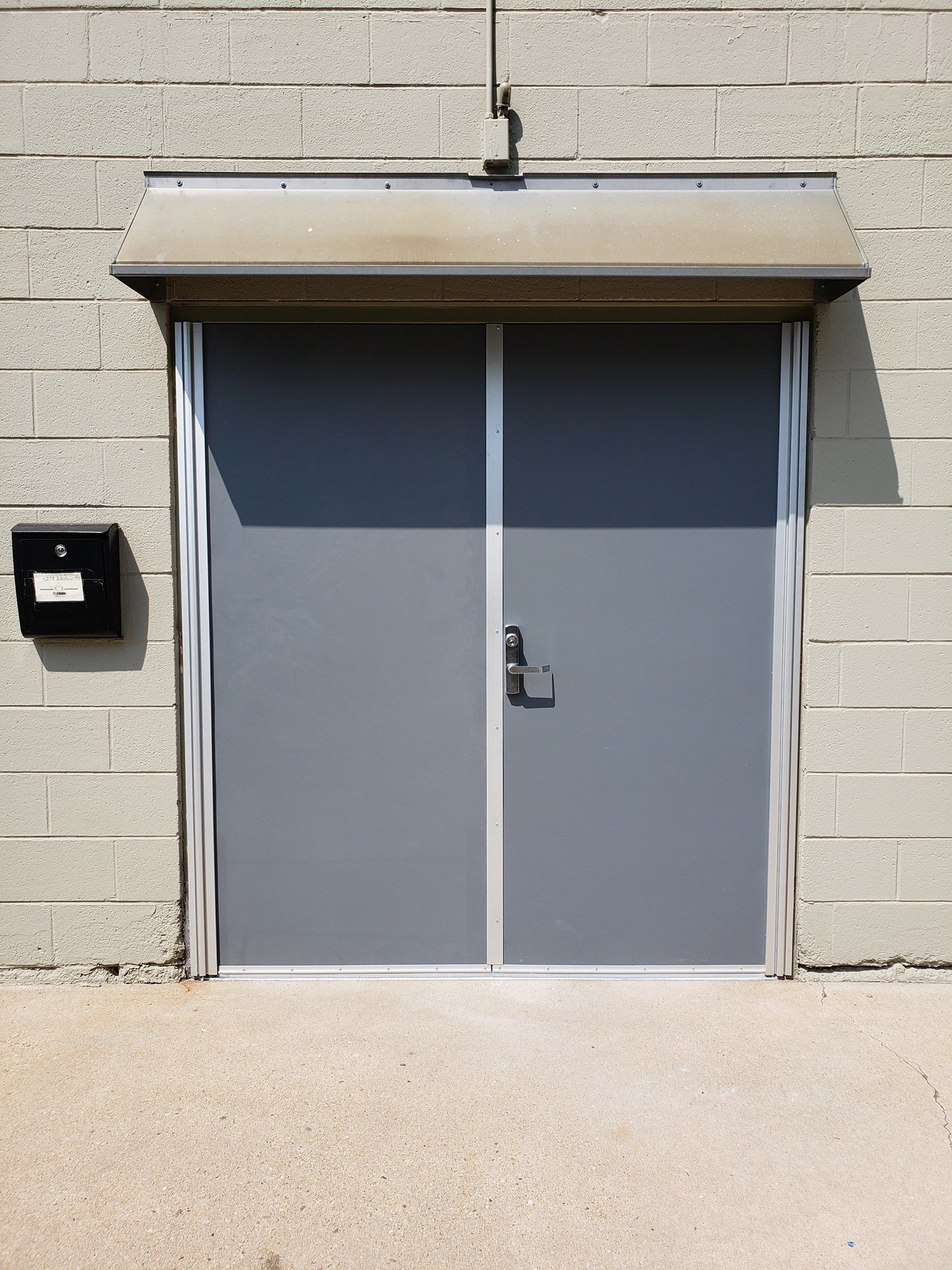 Gray double doors with metal trim on a cinder block building. A mailbox is to the left, and a metal awning is above.