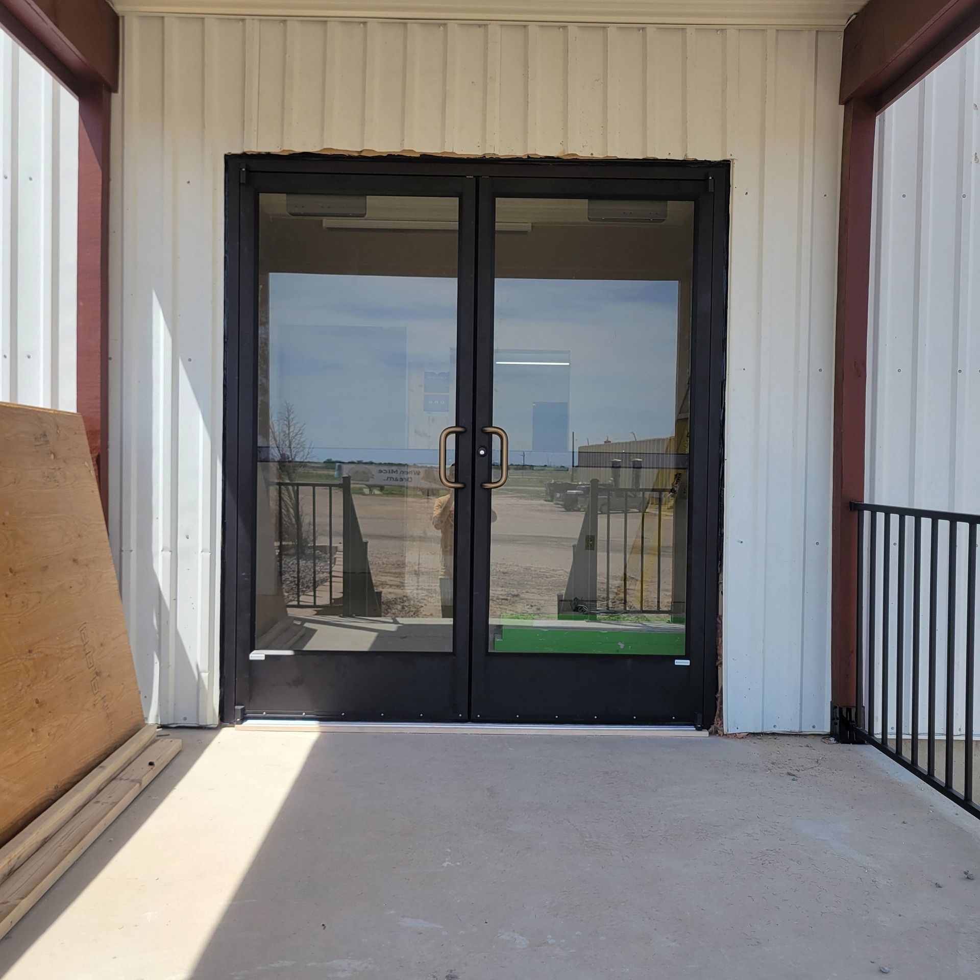 Double glass doors in a black frame, on a concrete porch, reflect a street and sky.