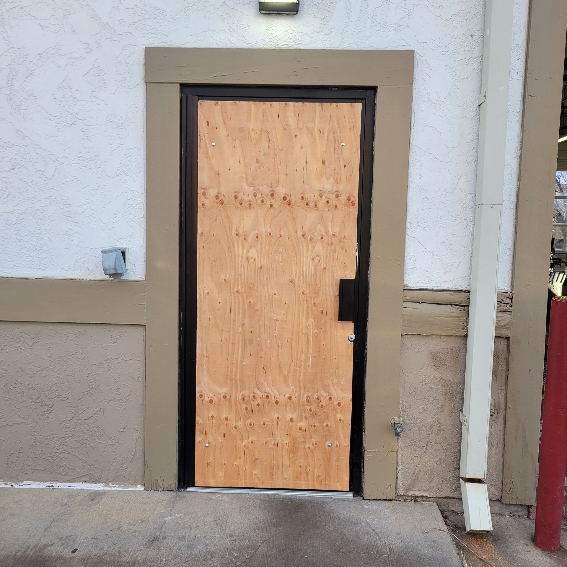 Boarded-up door on a beige and white building. Black frame around door, plywood secured with many small nails.
