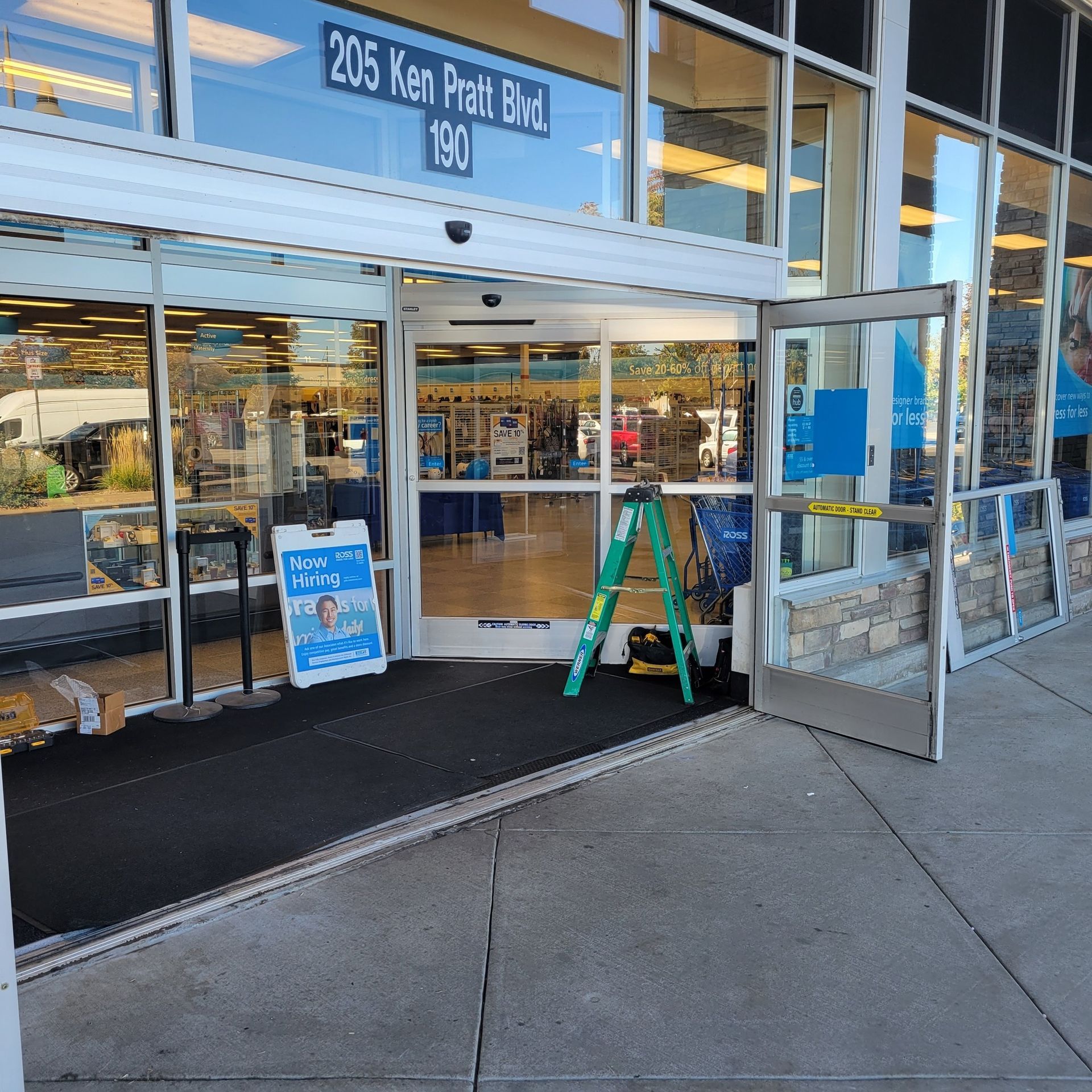 Entrance to a store with automatic doors; a ramp, a-frame sign, and a green ladder are present.