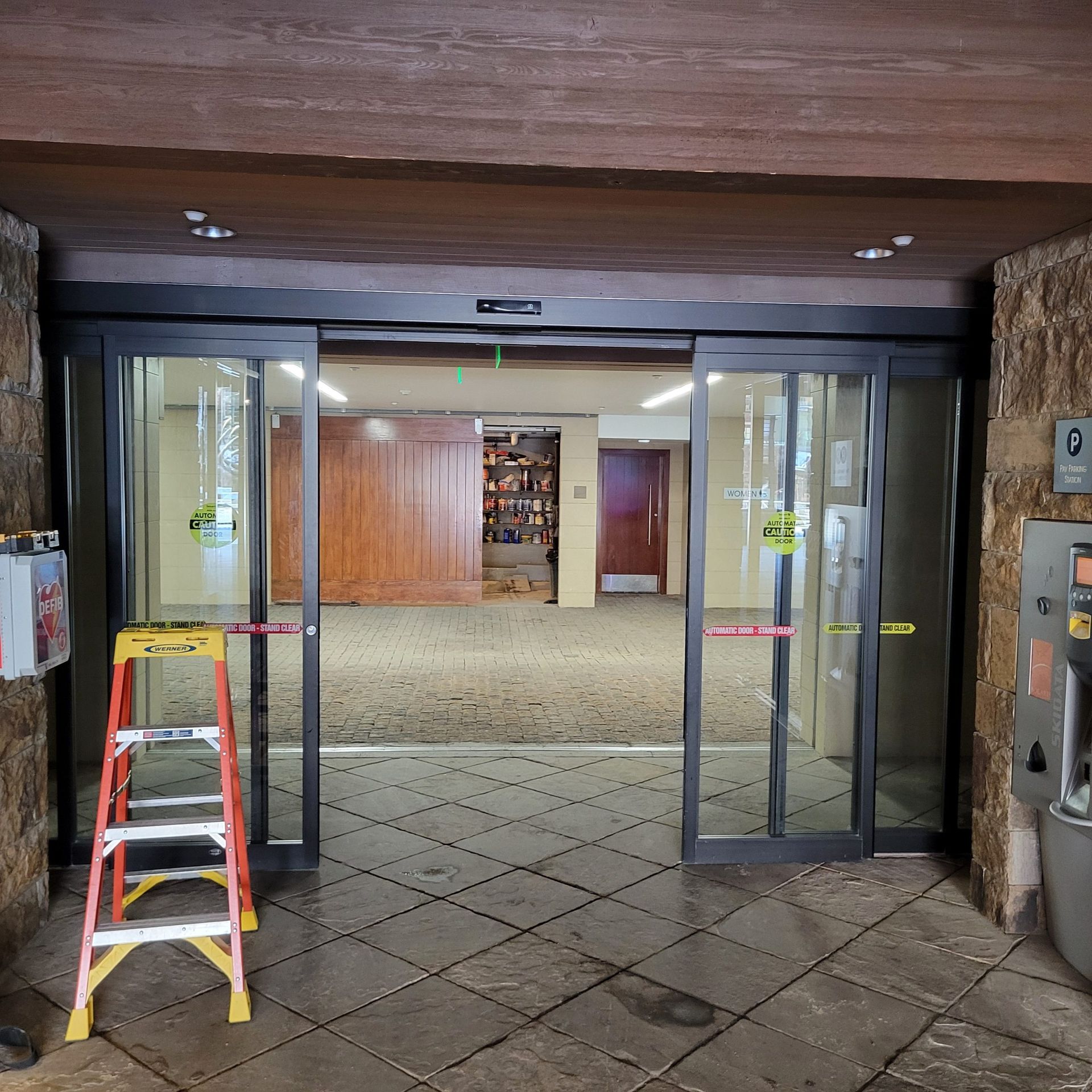 Automatic sliding glass doors open, revealing a hallway. A ladder stands in front of the doors.