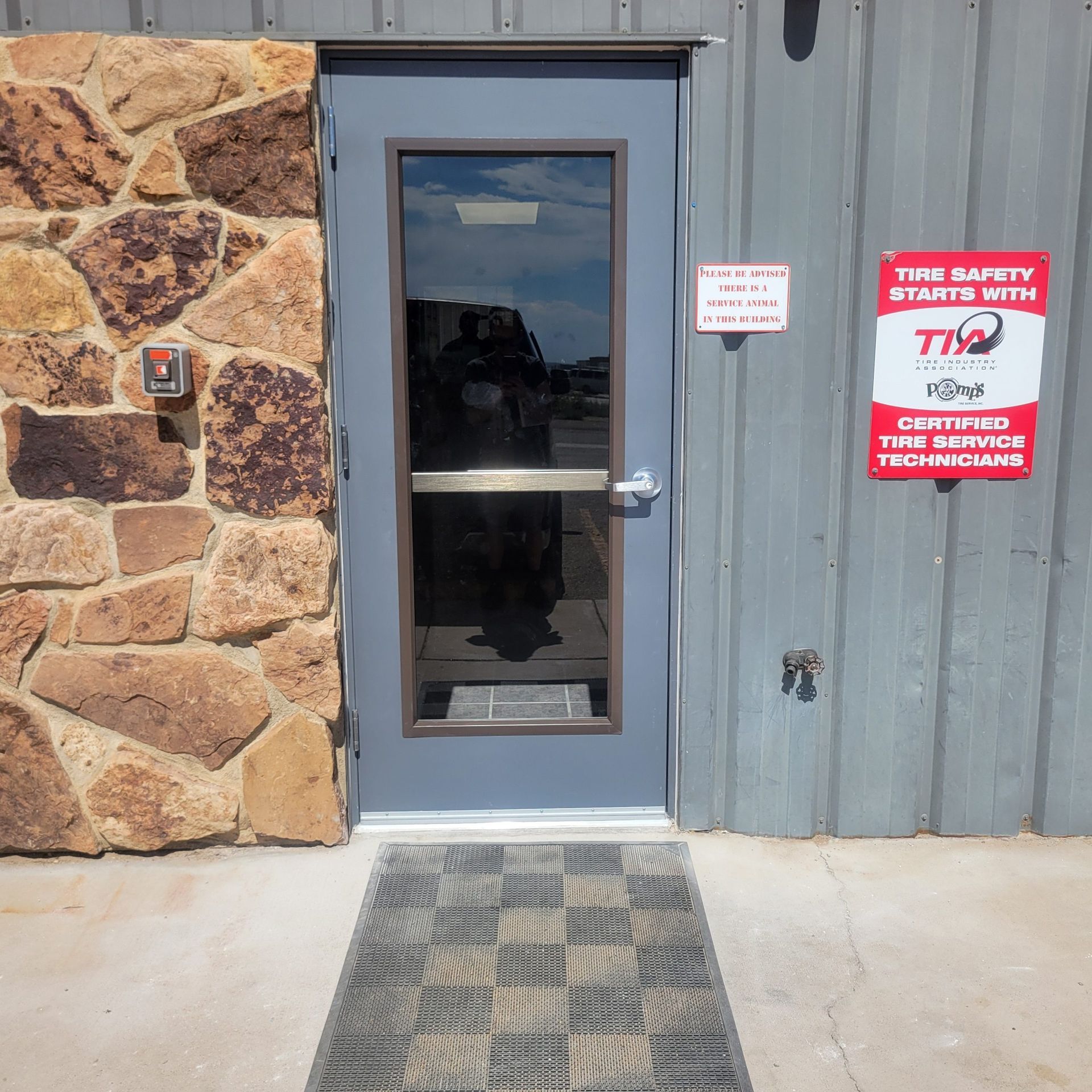 Doorway with stone wall and metal siding.  A doormat leads to a blue door with a window. Signage on the right.