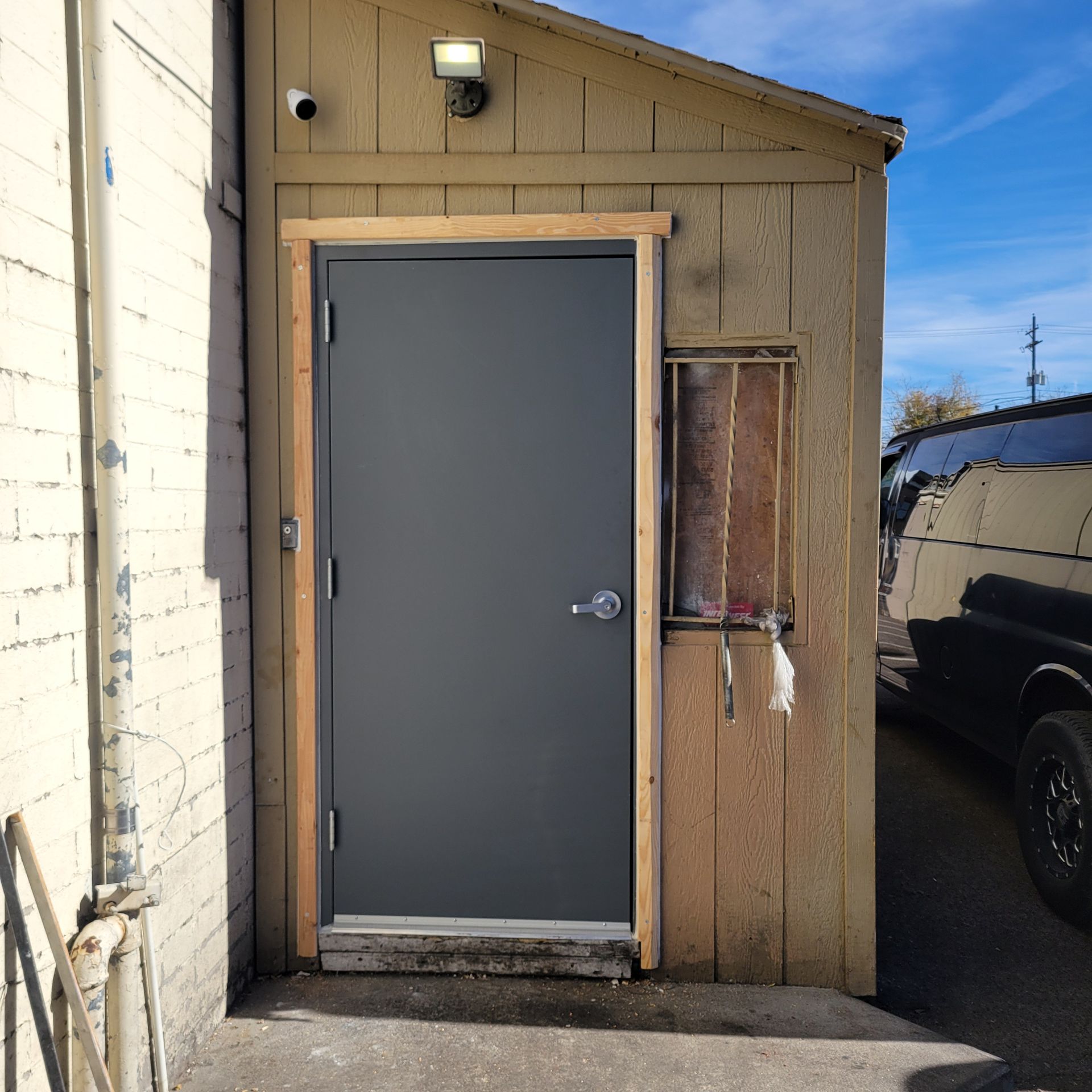 Gray door in a wooden building with a small window, security camera, and light. A black van is partially visible.