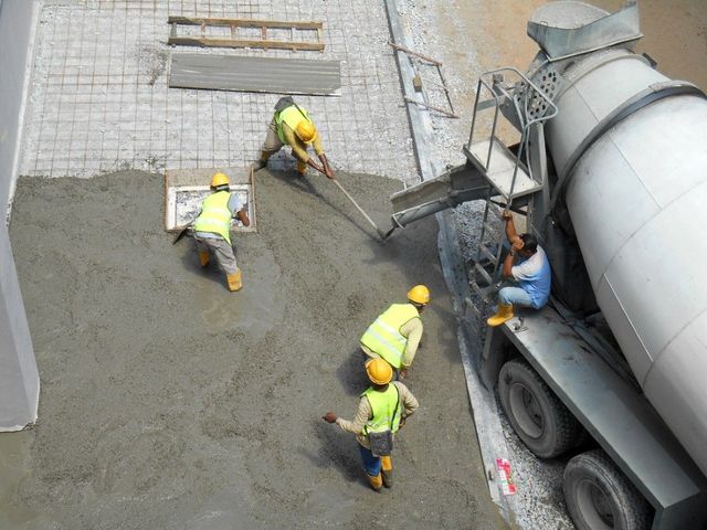 Four construction workers in neon vests and yellow hard hats pour and level wet concrete from a mixer truck.