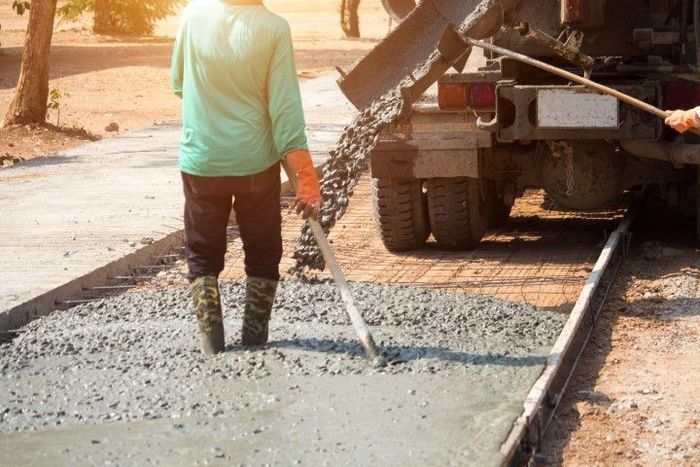 A construction worker in rubber boots uses a tool to spread wet concrete pouring from a cement truck onto a roadbed.