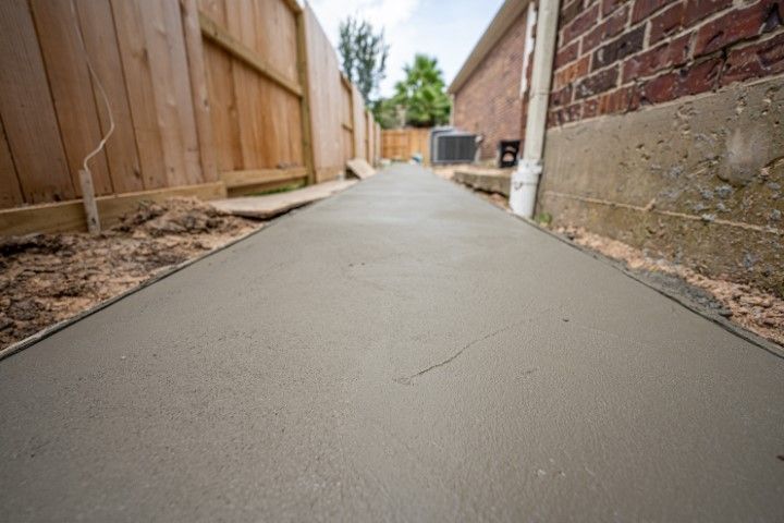 A freshly poured concrete sidewalk path between a wooden fence and a brick house wall.