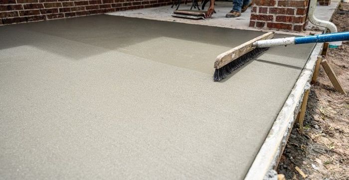 A concrete broom finish being applied to a freshly poured patio with a brick wall in the background.