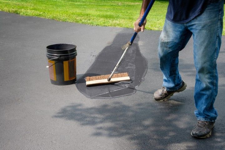 A person uses a long-handled brush to spread black asphalt sealant on a driveway next to a bucket of the material.