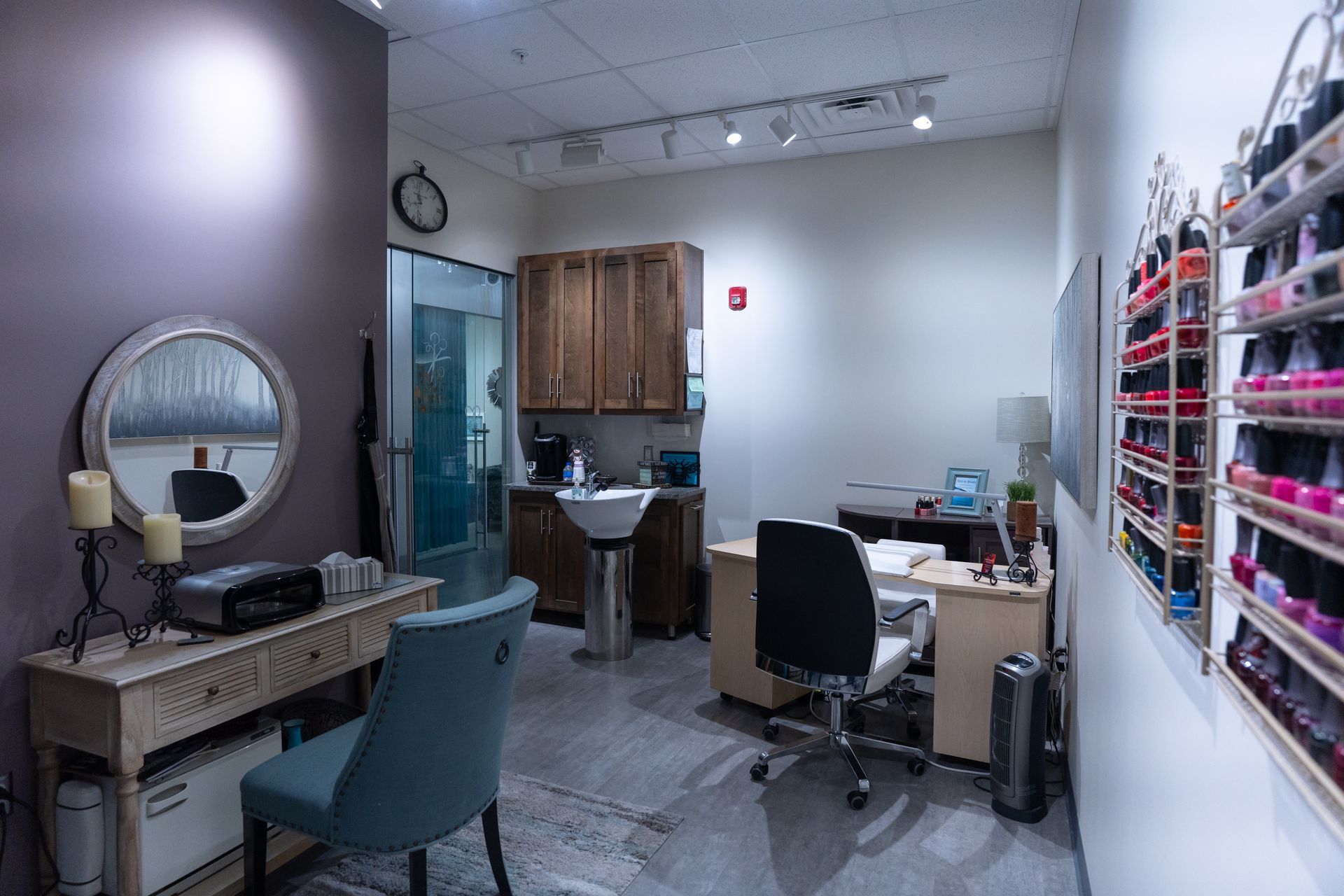 Nail salon interior with purple wall, manicure table, shelves of nail polish, mirror, and sink.