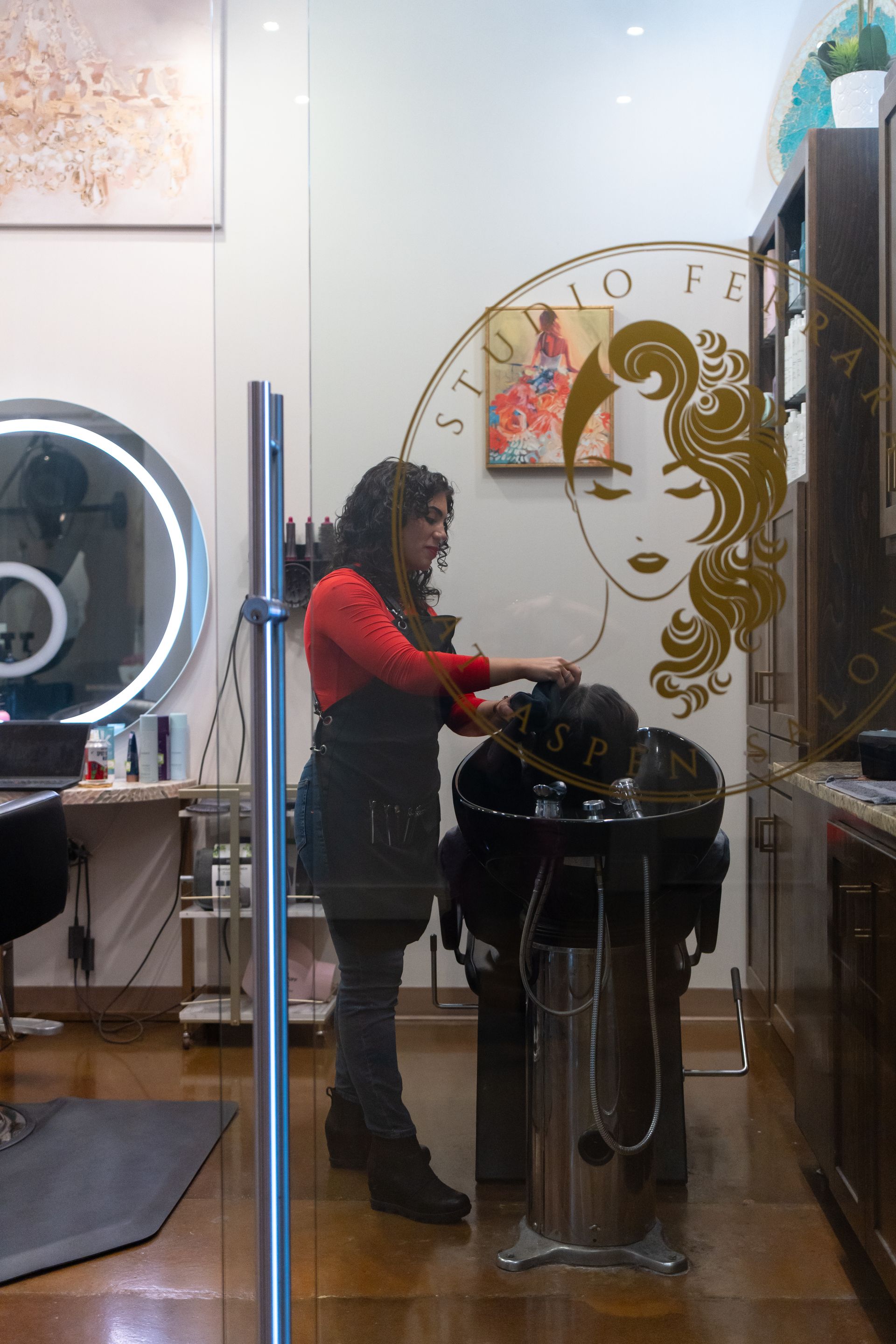 Woman in a red shirt and black apron, styling hair at a salon. Studio Fé design on window.