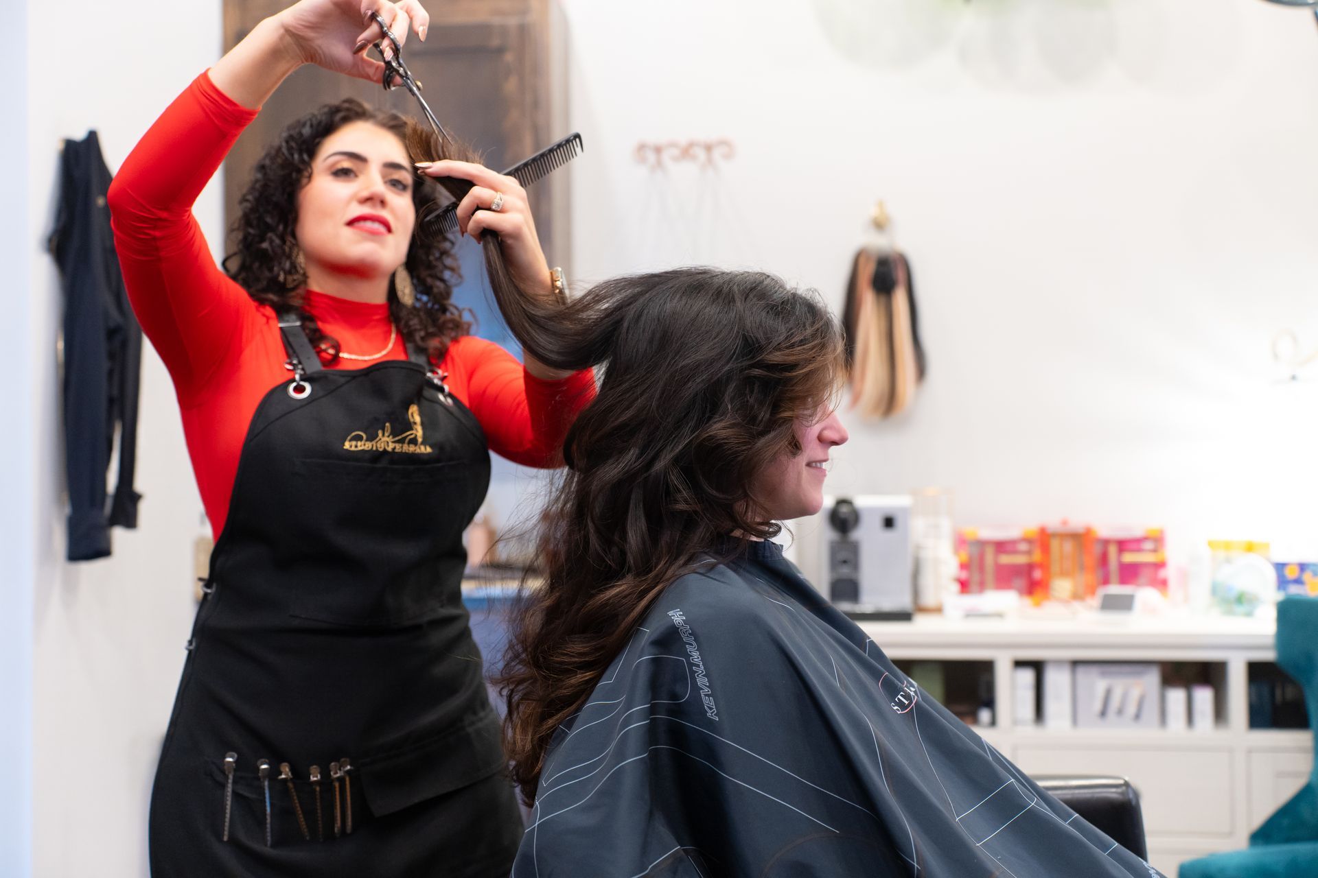 Hairdresser cutting a client's hair in a salon. The hairdresser wears a red shirt and black apron. Client sits wearing a cape.