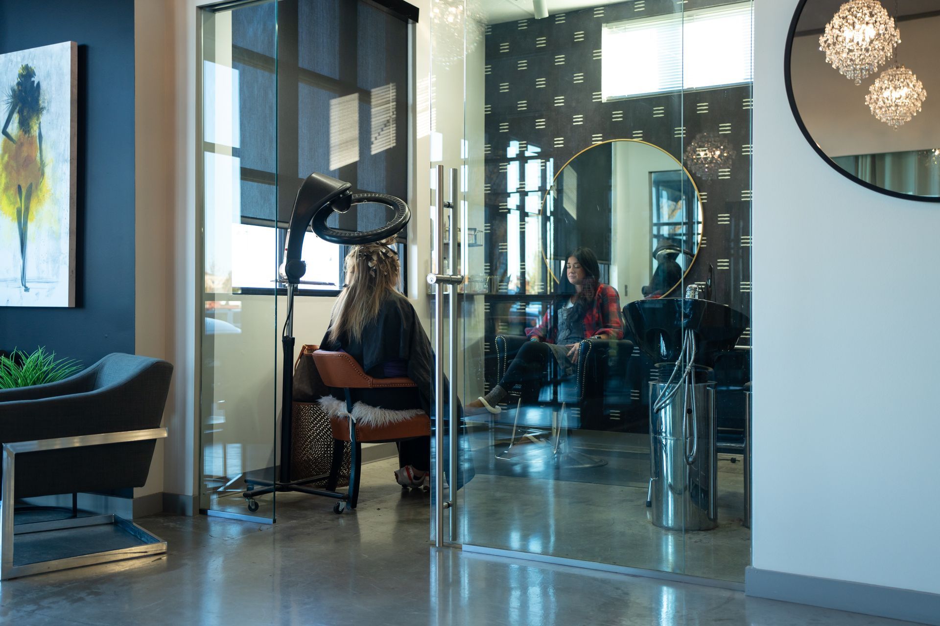A woman under a hair dryer in a salon. Through glass, another person sits at a styling station.