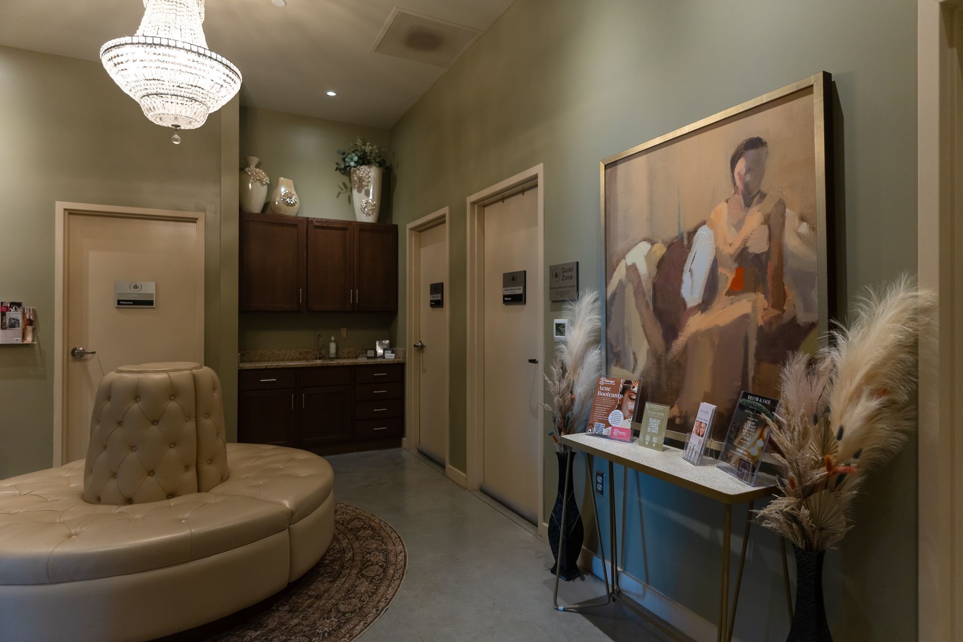 A waiting room with tan circular seating, artwork, and three closed doors. Green walls and a chandelier.