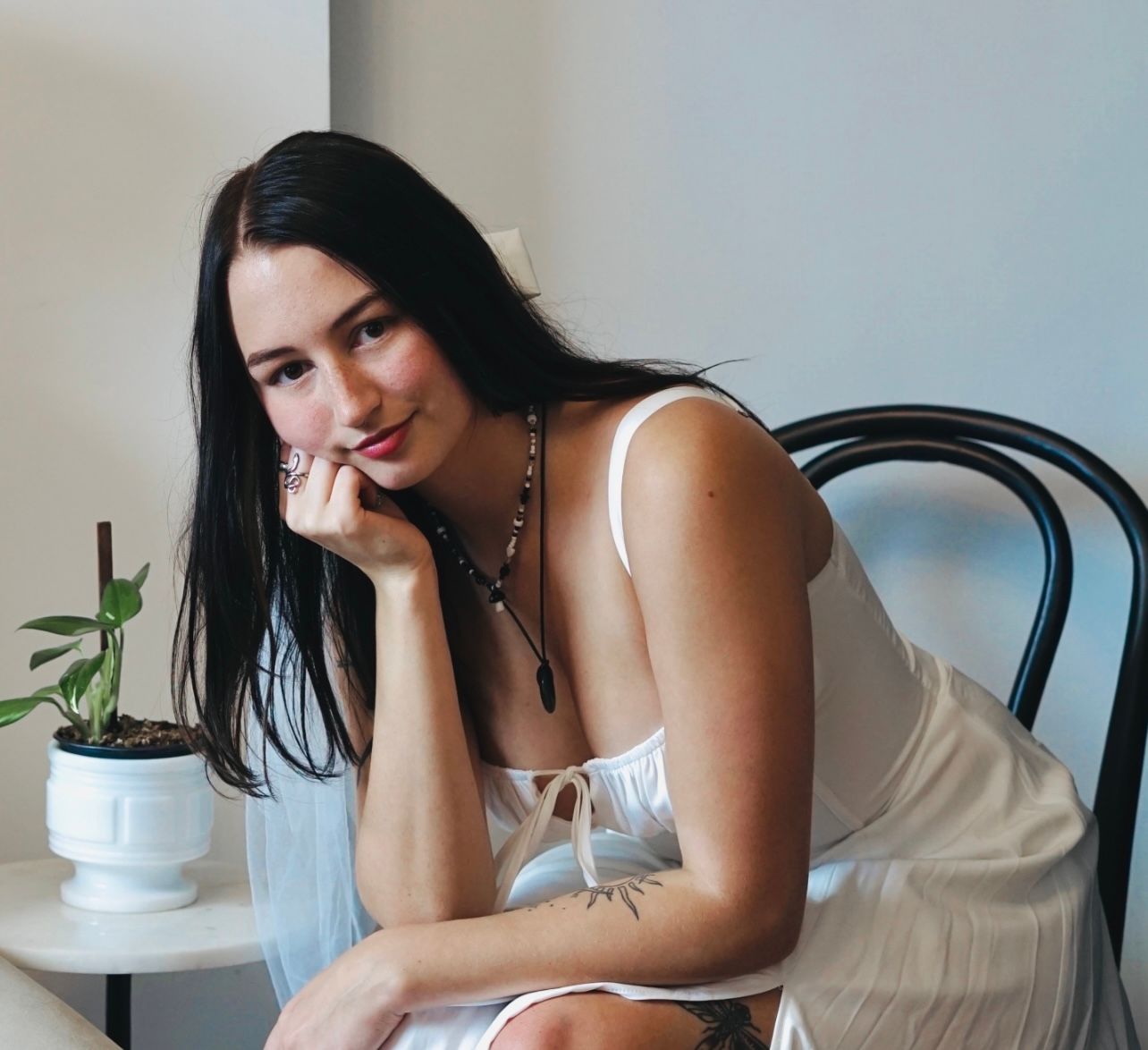 Woman in white slip dress, resting chin on hand, looking at viewer.