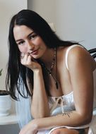 Woman in white slip dress, resting chin on hand, looking at viewer.