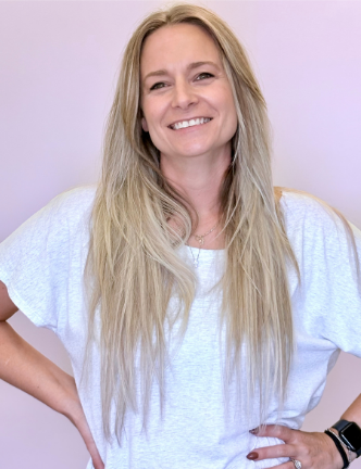Blonde woman smiling, hands on hips, wearing white shirt. Pink background.