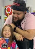 A barber and a smiling child, possibly at a salon, with the child in a cape and the barber wearing a hat.