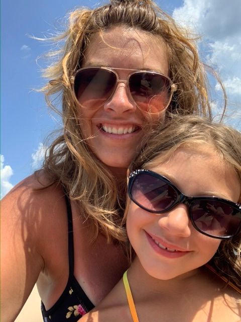 Woman and child smiling at the beach, wearing sunglasses, under a blue sky.