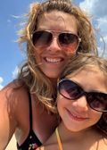 Woman and child smiling at the beach, wearing sunglasses, under a blue sky.