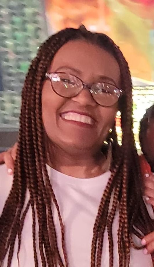 Woman with braided hair, glasses, and white top smiles. A blurred colorful background is behind her.