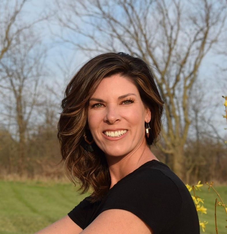Woman with brown hair smiling, wearing a black top, outdoors in front of trees.