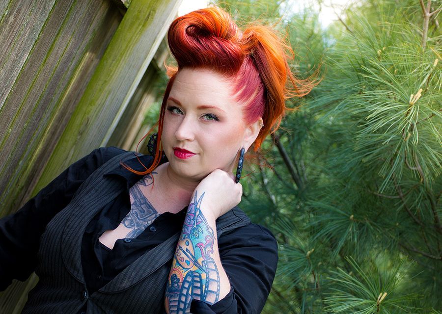 Woman with red hair and tattoos, resting chin on hand, wearing black clothing.