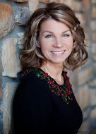 Woman with blonde hair smiles, leaning against a stone wall, wearing a black top with red floral embroidery.
