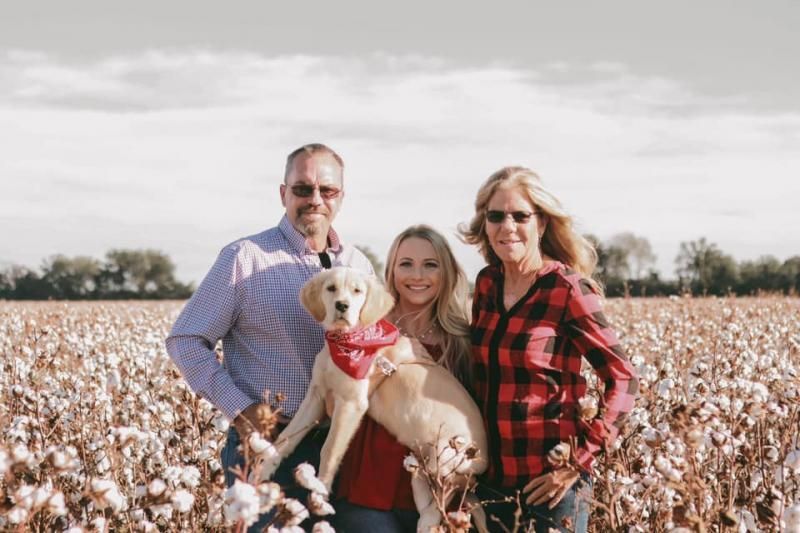 Three people and a golden puppy with a red bandana pose together in a field of blooming cotton under a bright sky.