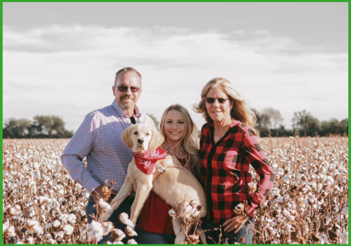 Three people and a light-colored dog in a red bandana pose together in a large, sunny cotton field.